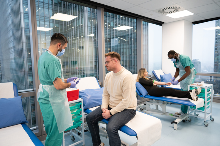 A patient sits on a bedside chair while a doctor holds a notepad and wearing scrubs.