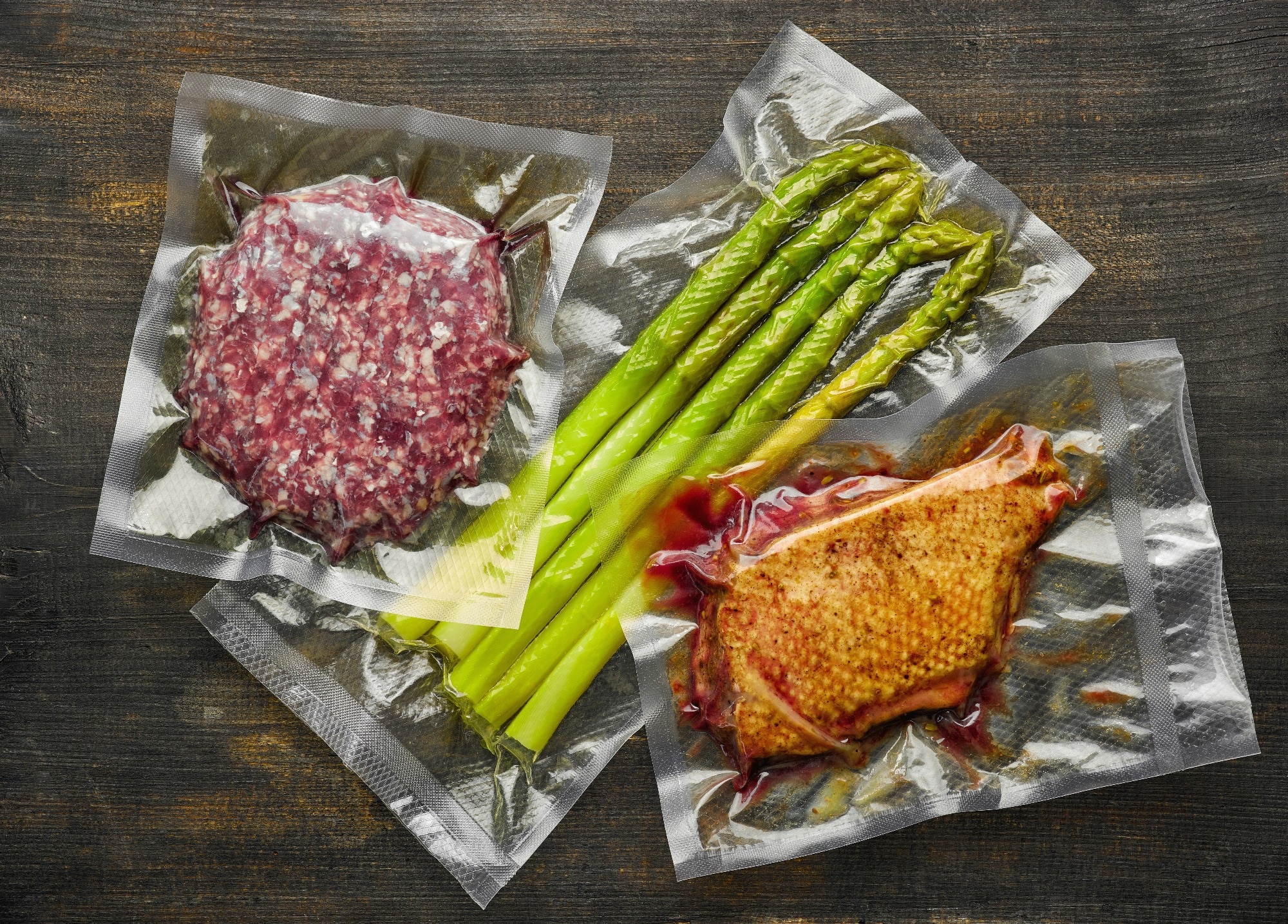 Duck breast, burger meat and asparagus vacuum sealed on wooden table